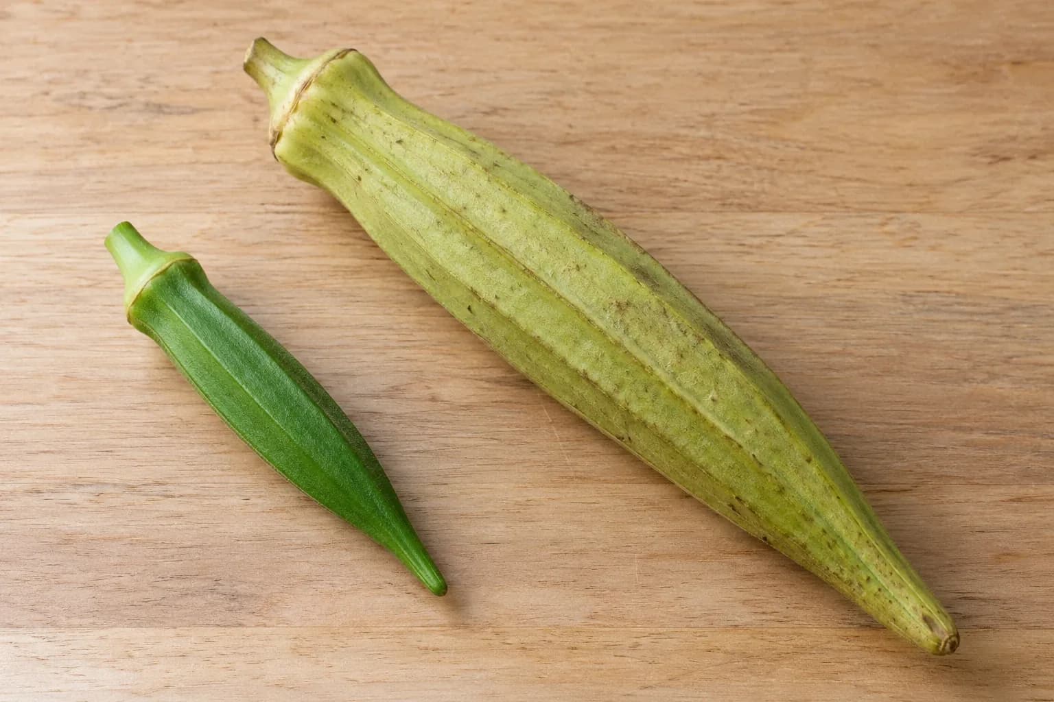 A small bright-green okra pod beside a much larger, paler overgrown pod on a wooden surface